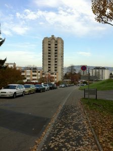 La Triemli Tower, ou la Hochhaus am Triemliplatz en allemand, vue depuis le haut d'une rue. La photo a été prise en automne. Des feuilles mortes se trouvent sur la chaussée.