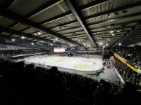 Photo en couleur de l'intérieur de la patinoire des Vernets à Genève pendant un match de hockey. Au premier plan, les gradins sont plongés dans le noir et le centre de la glace est éclairé. Plusieurs joueurs patinent sur la glace.