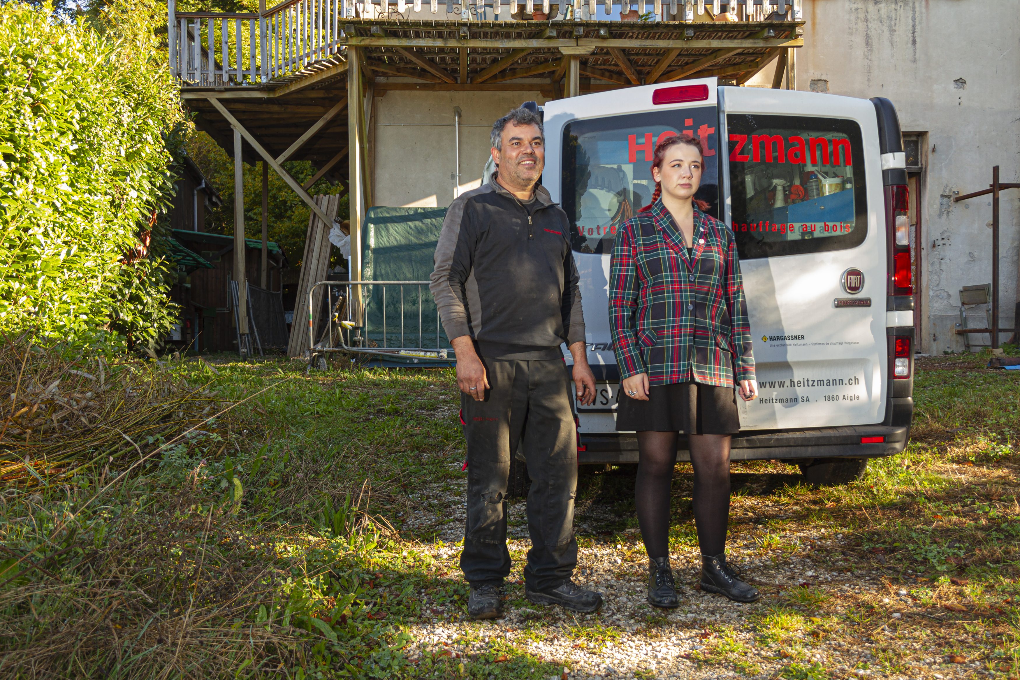 Un homme et une femme se tiennent debout dans la même position dans un jardin ensoleillé devant une camionnette de travail.