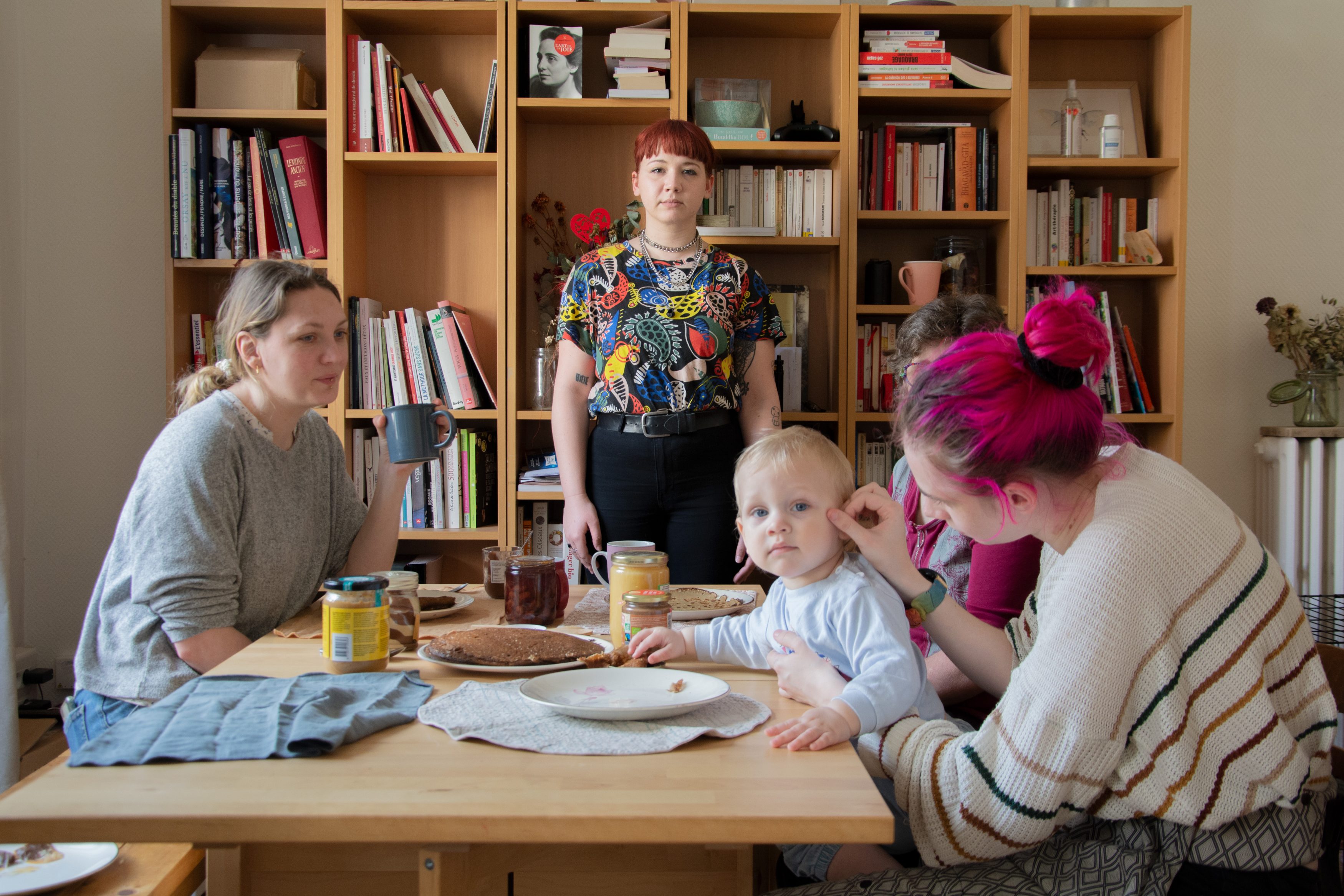 Une famille prend le goûter autour d'une table, au fond de la pièce, une femme se tient debout et fixe l'appareil photo